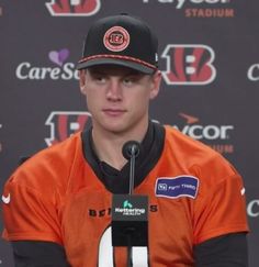 a man in an orange jersey and black hat sitting at a microphone with a baseball cap on his head