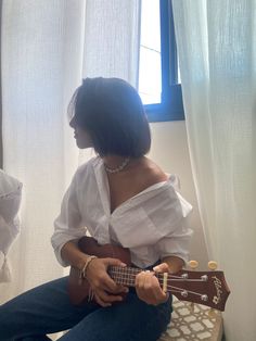 a woman in white shirt and jeans playing an ukulele on the floor next to a window