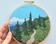 a hand holding a small embroidery hoop with a painting of a road in the woods