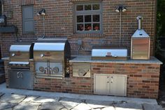 an outdoor kitchen with grills and sink in front of a brick building on a sunny day