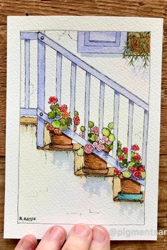 a hand is holding up a watercolor painting of flowers on the steps in front of a house