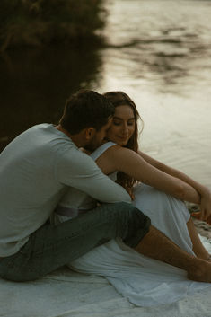 a man and woman sitting next to each other on the sand near water at sunset