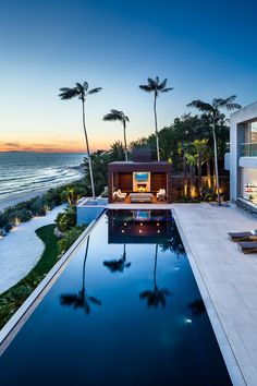 an outdoor swimming pool with lounge chairs and palm trees in front of the ocean at sunset