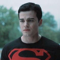 a young man wearing a superman t - shirt looking at the camera with trees in the background