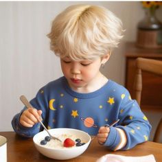 a little boy sitting at a table eating cereal with his spoon and fork in front of him