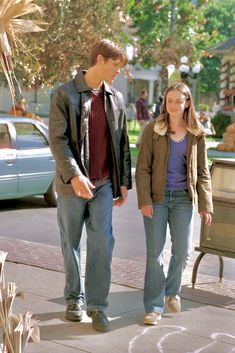 a man and woman standing next to each other in front of a bench on the sidewalk