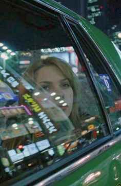 a woman sitting in the passenger seat of a green car at night with city lights behind her