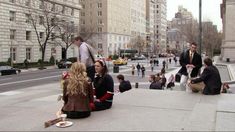 several people sitting on the ground in front of some tall buildings and one person is eating