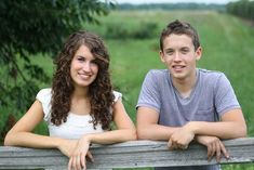 a boy and girl leaning on a wooden fence in front of a green grass field