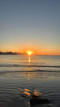 the sun is setting over the ocean with rocks in the foreground and mountains in the distance