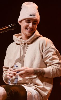 a young man wearing a white beanie smiles as he holds a microphone in front of him