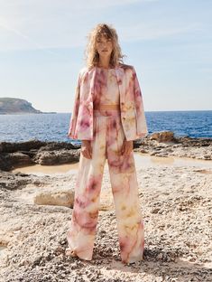 a woman standing on top of a sandy beach next to the ocean wearing a pink and yellow tie dye jumpsuit