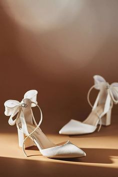 two pairs of white high heel shoes with bows on the toes and heels are shown in front of a brown background