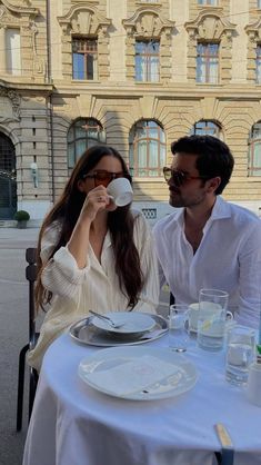 a man and woman sitting at a table drinking from paper cups in front of a building