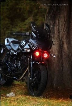 a motorcycle parked next to a tree in front of some grass and trees with its lights on