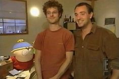 two men standing next to each other in front of a desk with a stuffed animal