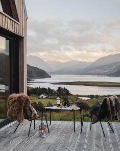 two chairs and a table on a wooden deck overlooking a lake with mountains in the background