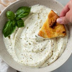 a hand holding a piece of pizza over a bowl of hummus