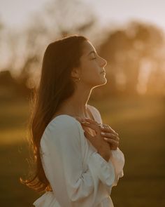 a woman standing in the grass with her hands folded and looking up at the sky