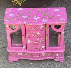 a pink dresser with flowers painted on the front and sides, next to a potted plant