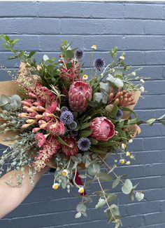 a person holding a bouquet of flowers in front of a blue brick wall with greenery