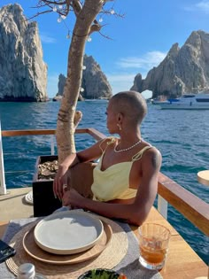 a woman sitting at a table with plates and drinks in front of some rocks on the water