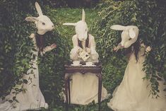 three women dressed up as rabbits sitting at a table in the middle of some bushes