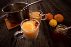 two mugs filled with liquid next to some oranges and a pot on a wooden table