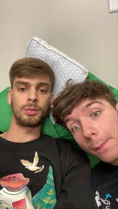 two young men laying next to each other on top of a green bed cover covered in pillows