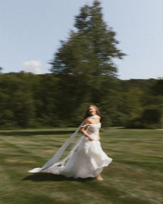 a woman in a wedding dress is walking through the grass with her veil blowing in the wind