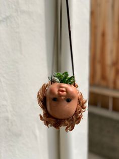 a doll head hanging from the side of a white wall with a plant in it's mouth