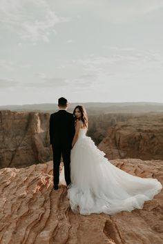 a bride and groom standing on the edge of a cliff looking out at the canyon