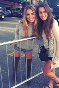 two beautiful young women standing next to each other in front of a metal fence and bus