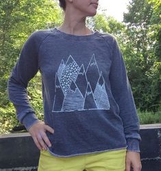 "Cozy, Soft, and Plush Sweatshirt is hand printed with my mountain design in white. this top is my go to as the weather starts to cool down at night. it keeps me cozy and stylish for all my outdoor activities. i wear this pullover headed to the gym, yoga, beach, camping adventure, hiking the trails and walking my dogs (+cat it's just the best top to wear for a little extra warmth and the perfect fall companion. this top is from alternative apparel. the material is a burnout french terry with an Winter Graphic Print Raglan Sleeve Tops, Graphic Print Raglan Sleeve Winter Top, Relaxed Fit Raglan Sleeve Sweatshirt With Graphic Print, Casual Winter Hiking Tops, Comfortable Graphic Print Winter Tops, Comfortable Winter Tops With Graphic Print, Comfortable Graphic Print Tops For Winter, Casual Relaxed Fit Tops For Hiking, Cozy Sweater With Graphic Print In Relaxed Fit