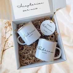 two white coffee mugs sitting in a box on top of a bed next to a note