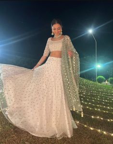 a woman in a white lehenga standing on grass at night with lights behind her