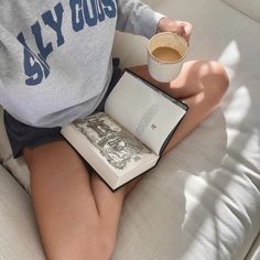 a person sitting on a couch holding an open book and cup of coffee