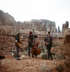 three men playing instruments in the desert near some rocks and trees with mountains in the background