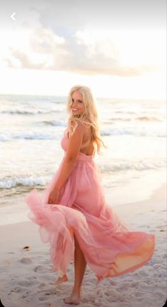 a beautiful blonde woman in a pink dress on the beach with her skirt blowing in the wind