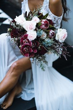 a woman in white dress holding a bouquet of flowers and greenery on her knee