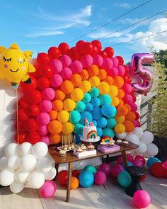 a table topped with lots of balloons next to a rainbow shaped cake and cupcakes