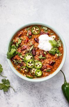 a bowl of chili with sour cream and jalapenos next to some cilantro