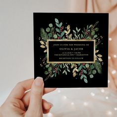 a person holding up a black and gold save the date card with greenery on it