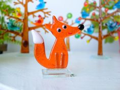 an orange glass fox figurine sitting on top of a white table next to trees