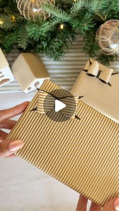 two hands holding a brown and white gift box in front of a christmas tree