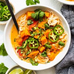 two bowls filled with noodles and vegetables on top of a white table next to limes