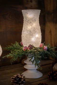 a white vase filled with pine cones and greenery next to a lit candle holder