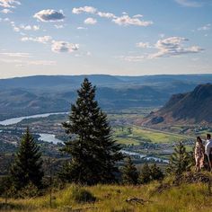 two people standing on top of a hill looking out at the valley and mountains in the distance