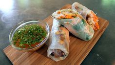 some food that is on a wooden board next to a small bowl and dipping sauce