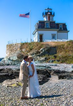 a bride and groom kissing on the beach in front of a light house with an american flag